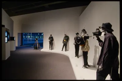 Five mannequins dressed in casual clothes and posed with surveillance gear line wall across from four tvs playing videos. 