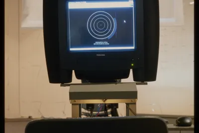 A black monitor displays concentric circles, and is mounted on a metal stand. A film screen is suspended in the background.