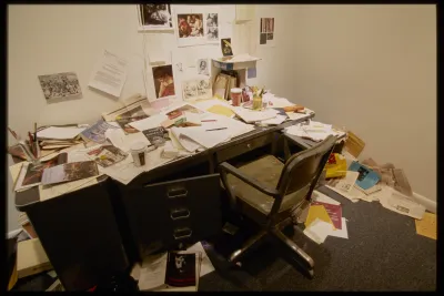 Desk and chair with disarray of art historian documents and office items covering the desk and spilling onto the floor.