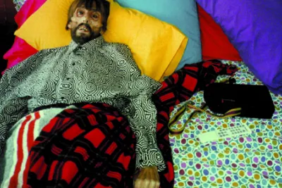  Photograph of bearded man with a sunken face on the edge of a bed covered in vibrant colored pillows and patterned sheets
