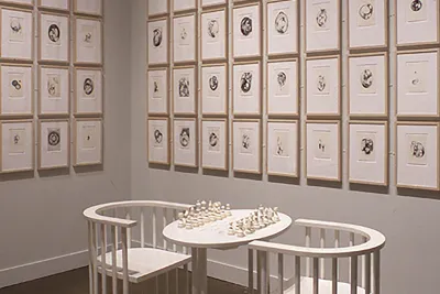 Installation of two white armchairs and a monochromatic chess table in front of a white wall, lined with framed prints 