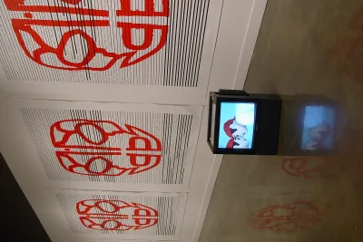 Covering the length of the gallery wall is a repeating image of a large red face-like design. A box monitor sits on the ground