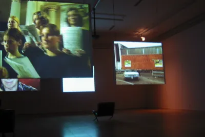 4 projection screens hang down from the ceiling in a dark room. One single chair sits empty in the background