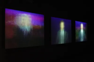 Three videos projected in a dark room show blurred images of men in suits in the middle of each frame.