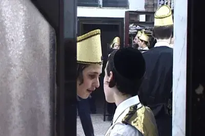 A young boy wearing a yarmulka watches from a doorway as carousing men dance in the street as part of the feast of Purim.