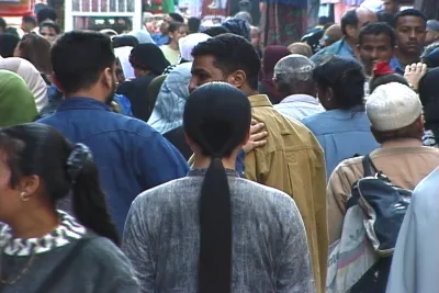 A woman with long black hair dressed in gray clothing stands with her back to the camera on a crowded street in Cairo.