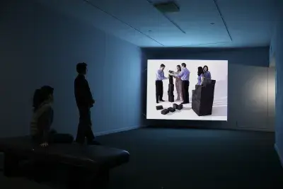 2 viewers looking at a large screen with 5 people working with blocks.