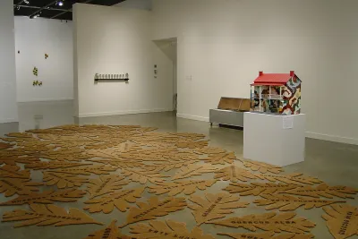 Leaf shaped stenciled cocoa mats grouped together and spread over gallery floor, next to house sculpture and other artworks.