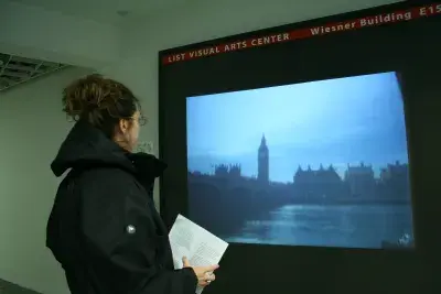 A visitor stands in front of a video projection in a hallway showing a view of a bridge over a river with buildings visible.