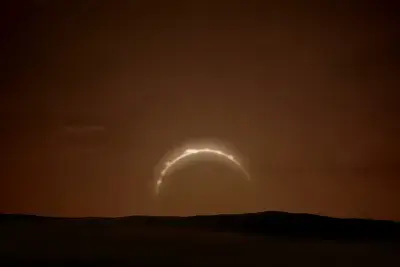 Amidst a total solar eclipse, a sliver of sunlight gives shape against a brown coloured sky with a dark shadowy landscape below.