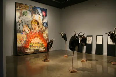 Large colorful painting of heads and fire facing 5 bestial horned masks on wood stakes with sketches on the right wall.