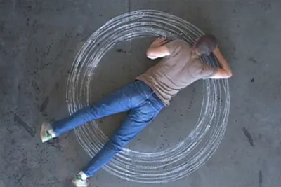 An aerial view of an artist stretching out on the floor with a piece of chalk. The artist draws concentric circles.