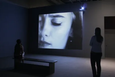 2 people in a dark gallery watching a video installation with a close shot of a woman's face.
