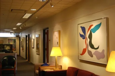 An entrance to a cosy office space with minimal abstract prints hung on the wall above a couch and chair with lamps beside.