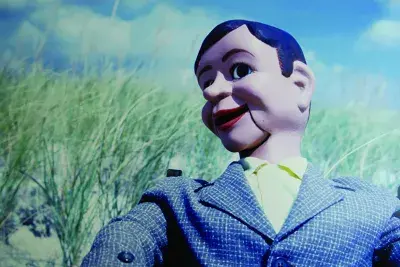 A close photograph of a ventriloquist dummy with wide eyes, smiling red lips, dressed in a suit jacket on a beach.