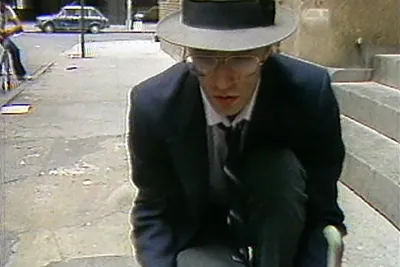 A video still shows a close view of a man dressed in a suit, tie, fedora hat and glasses kneeling on a city sidewalk.
