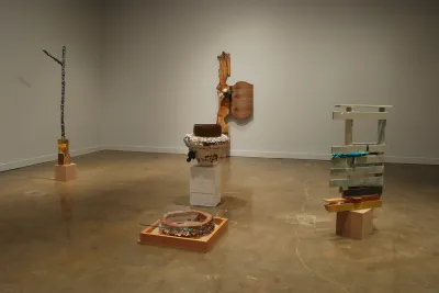 Five sculptures; three on low plinths, one leaning on a wall, and a flat, low sculpture with wood, a round object, and glass