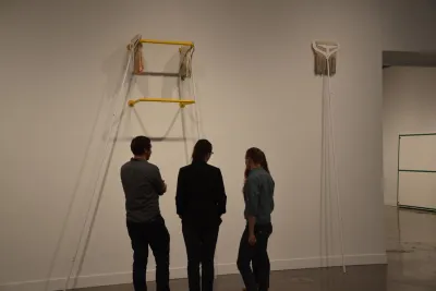 Three gallery visitors look up at a sculpture made of mops and yellow beams.