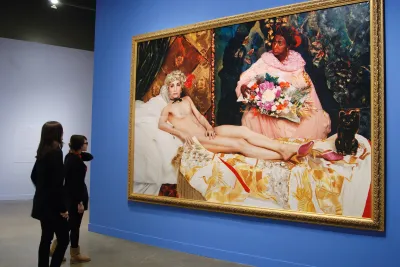 2 visitors view a large photo in a gold frame of a man portraying a nude woman lounging on a bed and a woman beside holding flowers. 