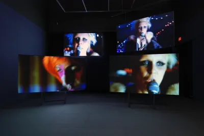 A dark room with four large video screens three of which show a rock singer in a music club from different angles.