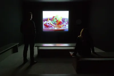 Two people stand in a dark gallery and view a projection of a closeup of pink food and lemon peel.
