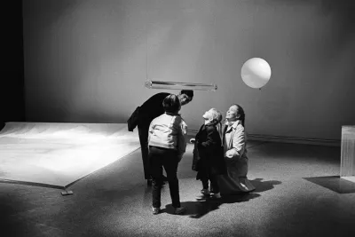 A photo of visitors viewing a clear cylindrical sculpture suspended in air from underneath it in Hans Haacke’s 1967 exhibition.