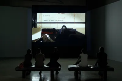 5 people sitting on benches in a dark gallery looking at a large movie screen with a still of a close up of a typewriter.