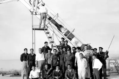 A black and white photo of people posing in front of a rocket. Two people are on a step ladder adjusting the rocket.