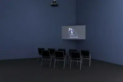 A dimly lit gallery space includes chairs and a screen with someone speaking at a podium that reads &quot;Walker Art Center&quot;.