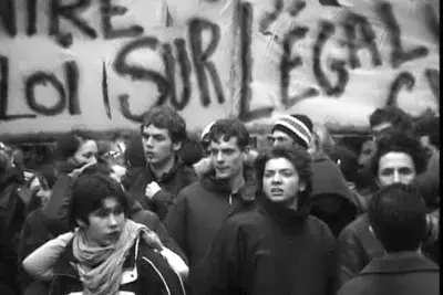 Black and white photo of several young people gathering in protest, a large handwritten sign filling the background.