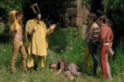 A man in a tall cylindrical hat and a gold tunic talks to 3 young men as they stand over a young man asleep in the grass.