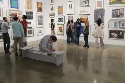 People crowd around gallery walls to view artwork and one person is taking notes on the bench.