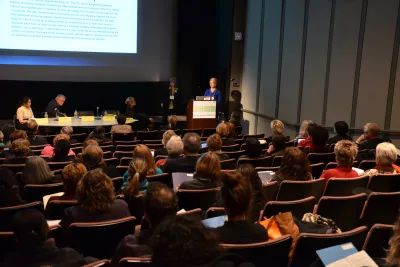 An audience seated in an auditorium turn to a speaker in a blue suit.