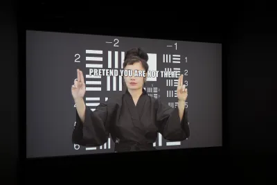 White text “PRETEND YOU ARE NOT THERE” covers the eyes of a woman in black kimono standing before black and white graphic.