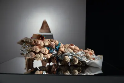 A cascade of tan snail shells on twisted aluminum foil and white fabric rests on a glassy-surface pedestal.