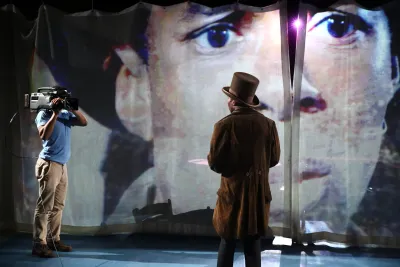 A camera person and a performer in a bown suit with a brown top hat stand in front of a projection of a face.