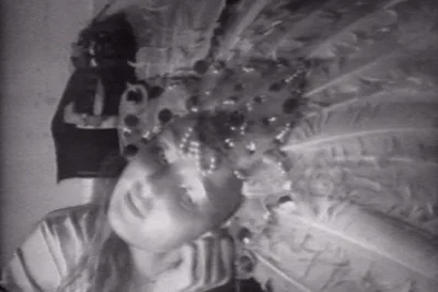 Black and white image of a female stage performer wearing a feather crown headdress with sequins and jewels. Her head is tilted to the side.