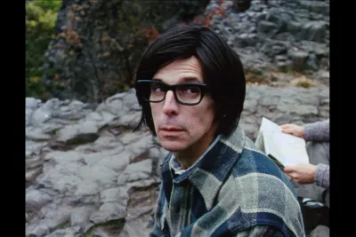 A close up of a person with dark hair and dark glasses wearing a plaid shirt. A set of hands holding a map is to the right.