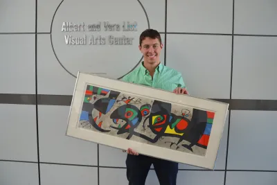 A man poses with an artwork next to a sign that reads " Albert and Vera List Visual Arts Center"