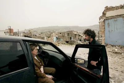 A man leans on the open passenger seat door of a black car and a woman is seated. The ruins of a city are pictured behind them.