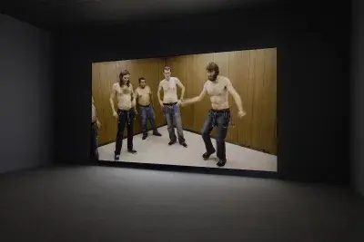 A large projection of four men shirtless people in denim jeans is situated in a dark gallery.