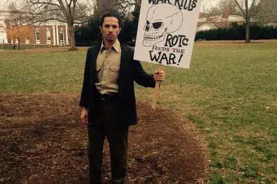 A man in a suit stands with a protest sign that reads "War Kills. ROTC feeds the war!"