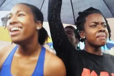 Three women stand under a black umbrella at a protest.