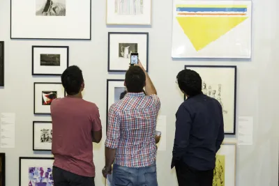 Three people view artwork on the gallery walls and one person in the center is taking a photo of the artwork.