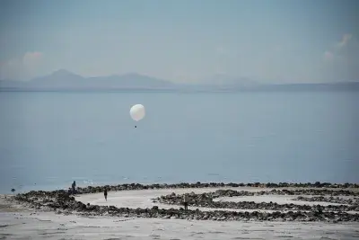 A white balloon floats over a spiral jetty, beach, and ocean.
