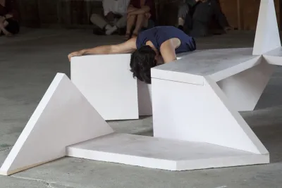 A performer in dark clothes stretches over a sculpture of white geometric shapes.