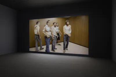 In a wood paneled space, five men looking toward the right, all wearing pants, tin foil vests, and belts with bells dangling