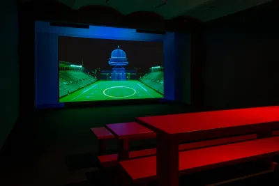 A gallery space includes a screen with an artwork featuring a soccer field in front of the Capitol building.