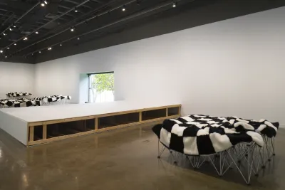 A black and white checkered fur blanket sits on multiple stilt structures in a gallery space.