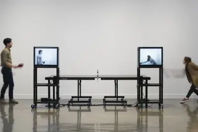 2 people playing ping pong at a table flanked by a pair of monitors, displaying players engaged in a simultaneous game. 
